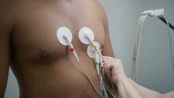 女性の心臓科医は 心臓病患者をホルターモニタリングするためのセンサーを置きます 医療センターの心臓病室での電気変動または心拍数測定中にアフリカ系アメリカ人男性 — ストック動画