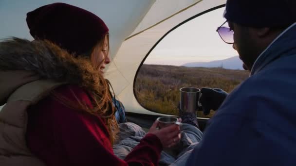 アフリカ系アメリカ人男性が白人女性とキスをする 複数のハイカーが話し お茶でカップを飲み 日没を見て 丘の上のテントに座っています 観光客は休暇中に山に休んでいる — ストック動画