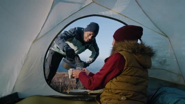 女人坐在帐篷里 手里拿着两杯茶 拿着沸腾水壶的非裔美国人在杯子里倒水 高山上的多族背包客家庭在寒冷多风的天气里热身 — 图库视频影像