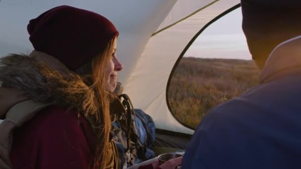 年轻的女游客和丈夫坐在山顶的帐篷里 白人女人手里拿着杯子 看着夕阳 与非裔美国男人交谈 一对夫妇在山上度假 — 图库视频影像