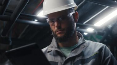 Endüstriyel imalat fabrikasında dijital tablet bilgisayar kullanan profesyonel Kafkasyalı mühendisi kapatın. Ağır endüstri uzmanları güvenlik üniforması, gözlük ve şapka modern fabrikada çalışıyor..