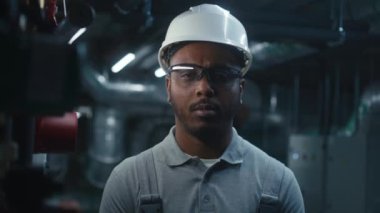 Afrika kökenli Amerikalı ağır sanayi işçisi iş yerinin yanında duruyor ve kameraya bakıyor. Modern endüstriyel fabrika, fabrika ya da enerji tesisinde güvenlik üniforması ve şapka mühendisi olarak çalışıyor..