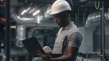 Erkek Afrika kökenli Amerikalı müfettiş dizüstü bilgisayarla boru hattını kontrol ediyor ve kameraya bakıyor. Çeşitli profesyonel teknisyen ekipleri modern fabrika üzerinde çalışıyor. Ağır endüstri konsepti. Portre.