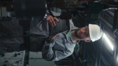 Güvenlik üniforması giyen ve şapka takan erkek ağır sanayi işçisi dijital tablet bilgisayarda çalışıyor. Profesyonel mühendis modern imalat fabrikası veya sanayi tesisi kurar. Dikey görünüm.