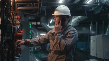 Güvenlik üniforması giyen, koruyucu gözlük takan ve baret takan erkek ağır sanayi işçisi boru hattı sistemini denetliyor, telefonla konuşuyor. Mühendis modern imalat fabrikası veya sanayi tesisi işletiyor.