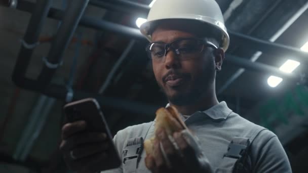 非裔美国工程师吃三明治 然后打电话上网 在工业工厂或能源设施工作的专业工人 头戴制服和防护硬礼帽 休息休息 靠近点 — 图库视频影像