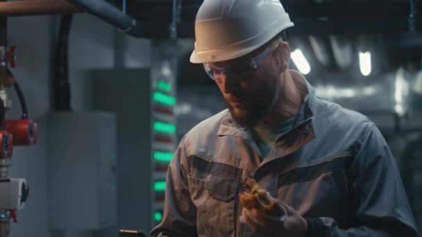 穿着安全制服 护目镜和硬礼帽的男性重工业工人吃汉堡包并使用电话 在现代制造厂或工业设施休息的专业工程师 — 图库视频影像