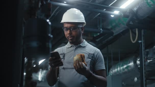 非裔美国人技师吃三明治 用手机上网 在现代工业工厂工作的安全制服和防护硬礼帽专业工程师 — 图库视频影像