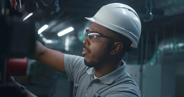Close up of African American heavy industry worker in safety uniform and hard hat inspecting and adjusting energy system. Professional engineer works on modern manufacturing or industrial plant.