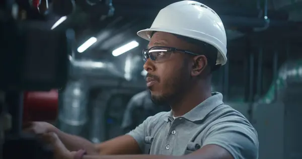 Close up of African American heavy industry worker in safety uniform and hard hat inspecting and adjusting energy system. Professional engineer works on modern manufacturing or industrial plant.