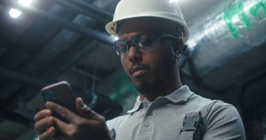 Tatil sırasında telefon kullanarak internette sörf yapan Afrikalı Amerikalı mühendisi kapatın. Güvenlik üniforması ve şapka giyen ağır sanayi işçisi modern imalat fabrikası veya sanayi fabrikasında çalışıyor..