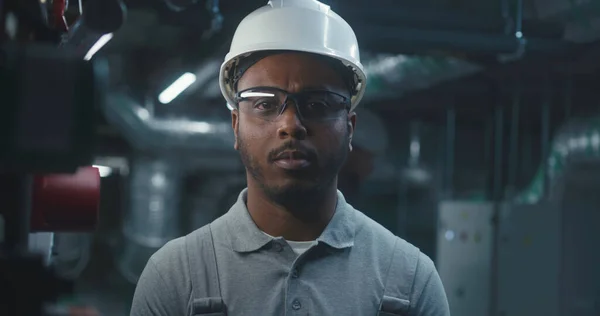 African American heavy industry worker stands near workplace and looks at camera. Professional engineer in safety uniform and hard hat works at modern industrial factory, plant or energy facility.