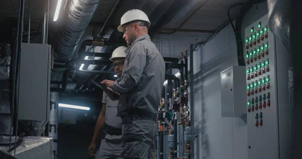 Professional Caucasian engineer in safety uniform, goggles and hard hat uses digital tablet computer, adjusts energy system on modern factory. Technician worker talks with African American colleague.