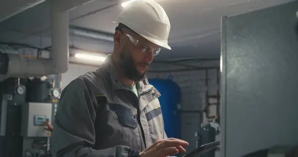 Professional Caucasian engineer in safety uniform and protective hard hat uses digital tablet computer to check and adjust energy system. Industrial worker works on modern manufacturing factory.