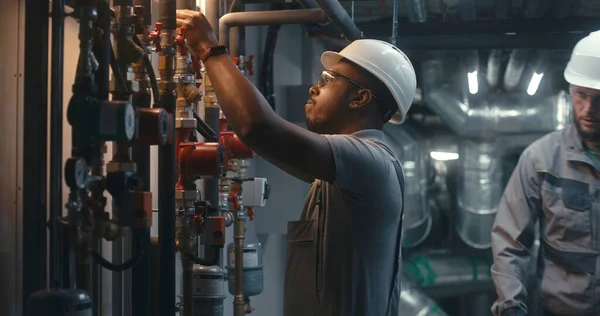 African American heavy industry worker wearing safety uniform and hard hat inspects piping system on modern plant or industrial energy facility. Professional engineer maintains manufacturing factory.