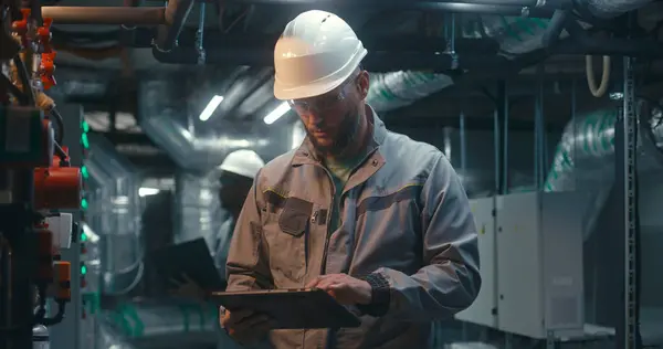 Diverse team of factory specialists work on modern plant using laptop and tablet computers. Industrial workers wearing safety uniform and hard hat inspect pipeline system. Heavy industry concept.