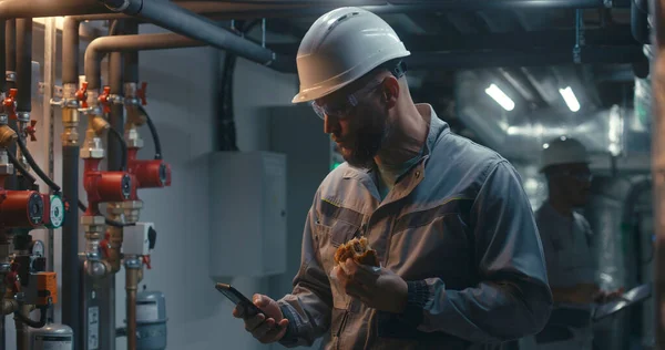 Professional engineer works at modern factory, eats burger and uses phone. In background African American colleague inspects pipe system using tablet computer. Industrial manufacturing plant concept.
