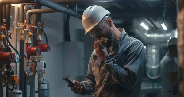 Professional engineer works at modern factory, eats burger and uses phone. In background African American colleague inspects pipe system using tablet computer. Industrial manufacturing plant concept.