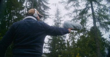 Kulaklık takan ses tasarımcısı, kozalaklı ormanda film için doğanın sesini kaydediyor. Kürklü ön cam mikrofon kullanıyor. Beyaz adam dışarıda profesyonel ses ekipmanlarıyla çalışıyor. Yavaş çekim.