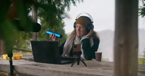 Caucasian woman in headphones plays shaker musical instrument, sits in gazebo in forest. Female musician creates and records music outdoors, uses laptop, phone on tripod, MIDI keyboard and microphone.