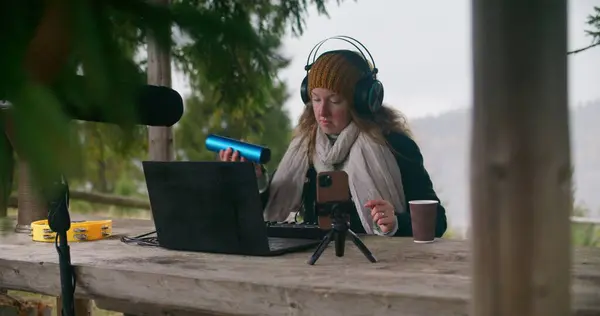 Caucasian woman in headphones plays shaker musical instrument, sits in gazebo in forest. Female musician creates and records music outdoors, uses laptop, phone on tripod, MIDI keyboard and microphone.