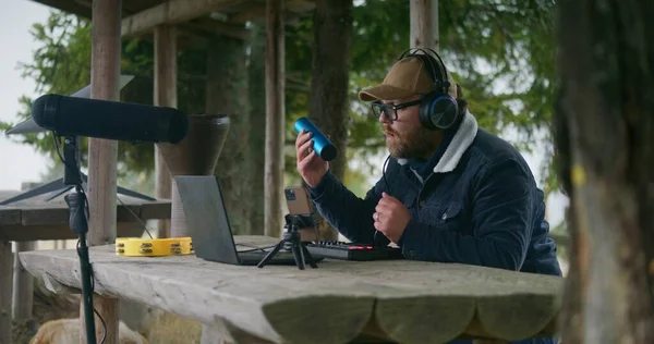 Caucasian musician in headphones sits in gazebo in forest, uses shaker and digital electric piano to create music. Man records song on phone on tripod and laptop during vacation trip to mountains.