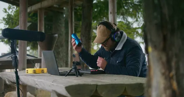 Caucasian musician in headphones sits in gazebo in forest, uses shaker and digital electric piano to create music. Man records song on phone on tripod and laptop during vacation trip to mountains.