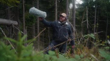 Kulaklık takan beyaz bir adam profesyonel ses ekipmanları kullanarak kozalaklı ağaçlarda film çekmek için doğanın sesini kaydediyor. Ses tasarımcısı dışarıda kürklü ön cam mikrofonuyla çalışıyor. Film yapımcılığı.