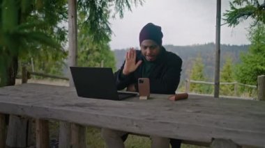 Afrika kökenli Amerikalı iş adamı ahşap çardakta oturur, online konferansta dizüstü bilgisayar ve telefon kullanarak konuşur. Turist güzel ormanda tatil sırasında müşterilerine uzaktan danışıyor. Açık hava işi..