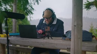 Kulaklıklı beyaz müzisyen çardakta dizüstü bilgisayarlı, telefon tripodlu, müzik aletli, şarkı besteleyip ritmik bir şekilde hareket ediyor. Yetişkin adam açık havada müzik yapmak için MIDI klavye kullanıyor.