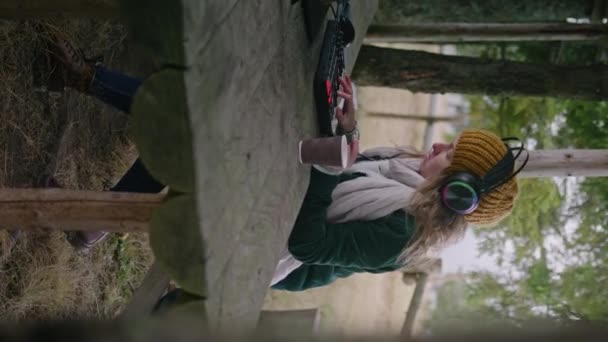屋外のMidiキーボードでヘッドフォンを着用している白人女性 女性ミュージシャンは 音楽を作成し レコーディングし 曲をミックスし 山林のガゼボに座っている間ラップトップを使用します 垂直ビュー — ストック動画