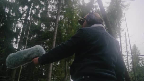 ヘッドフォンを着用している白人男性は プロのオーディオ機器を使用して複雑な木材で映画の自然の音を記録します サウンドテクニシャンは 屋外の毛皮風ジャマーマイクロフォンで動作します ローアングル — ストック動画
