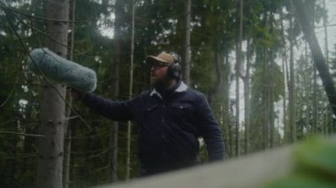 Kulaklık takan ses teknisyenleri kozalaklı ağaçlarda film için doğanın sesini profesyonel ses ekipmanları kullanarak kaydeder. Beyaz adam dışarıda kürklü rüzgâr engelleyici mikrofonla çalışıyor. Film yapımcılığı.