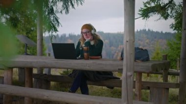Kulaklıklı yetişkin bir kadın ahşap çardakta oturur, müzik dinler ve dizüstü bilgisayar kullanarak internette sörf yapar. Gözlüklü turistler tatil sırasında güzel ormanda vakit geçirirler. Açık hava eğlencesi konsepti.