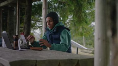 Afrika kökenli Amerikalı genç çocuk ahşap bir çardakta oturuyor, bilgisayar ve telefon kullanarak öğretmeniyle konuşuyor. Turist, dağ ormanlarında tatillerde uzaktan eğitim görüyor. Açık hava eğitim konsepti.