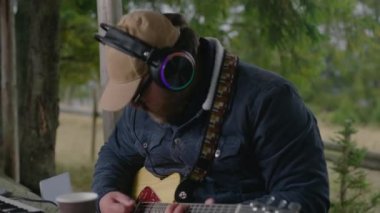 Kafkasyalı kulaklıklı adam gitar çalıyor ve ahşap çardakta oturmuş müzik kaydediyor. Profesyonel erkek müzisyen, besteci ormanda yeni bir şarkının ustası. Müzik oluşturma ve üretme.