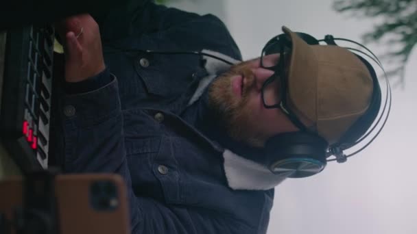 男歌手戴着耳机在Midi控制器上室外演奏 白人男子创作和记录音乐 使用笔记本电脑和电话在三脚架上 而坐在森林里的Gazebo 垂直录像 — 图库视频影像