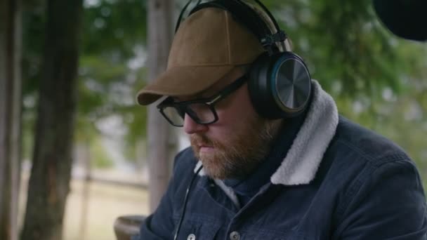 成年男性音乐家坐在木制凉亭中 创作新的作曲 在美丽的森林里度假时 戴着眼镜和耳机的游客可以远程工作 室外音乐制作概念 — 图库视频影像