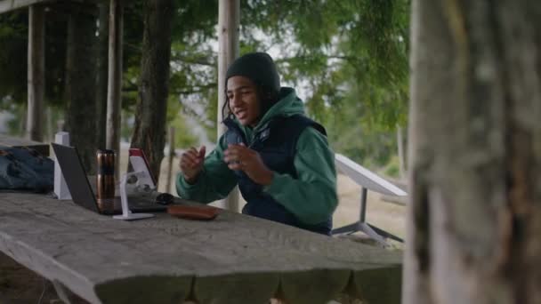 アフリカ系アメリカ人の少年が木製のガゼボに座り 三脚でラップトップと電話を使ってビデオ通話をする ティーンエイジャーは美しい山林で休暇中に遠隔的に研究しています アウトドア研究コンセプト — ストック動画