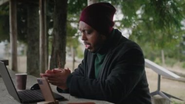 Afrika kökenli Amerikalı bir adam ahşap bir çardakta oturur ve tripodun üzerinde telefonla görüntülü konuşma yapar. Turist, güzel ormanda tatil sırasında çalışanlarla uzaktan konuşuyor. Açık hava işi kavramı.