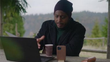 Afrika kökenli Amerikalı iş adamı ahşap çardakta oturur, kahve içer ve video görüşmesinde telefonla konuşur. Turist, güzel ormanda tatil sırasında çalışanlarla uzaktan konuşuyor. Açık hava iş kavramı.