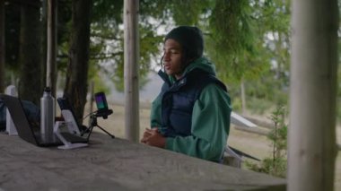 Afro-Amerikan flaması ahşap bir çardakta oturur ve profesyonel mikrofona konuşur. Genç, dağ ormanlarında tatil yaparken izleyiciler için içerik yaratmak için dizüstü bilgisayar ve telefon kullanıyor..