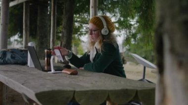 İş kadını ahşap çardakta oturur, online konferansta dizüstü bilgisayar ve telefon kullanarak konuşur. Kulaklıklı turist, güzel ormanda tatildeyken müşterilerine uzaktan danışıyor. Açık havada çalışmak.