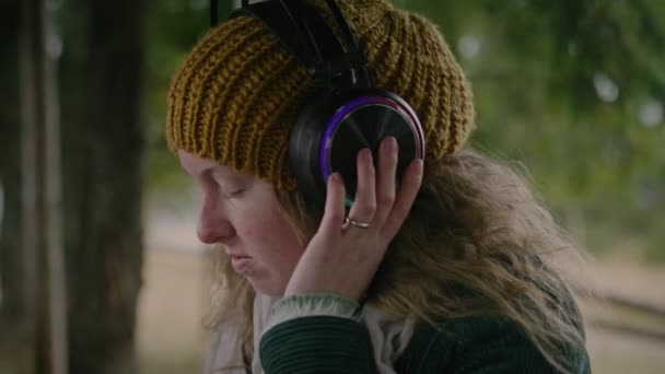 森のガゼボに座っているヘッドフォンで音楽を聴いている白人女性の近く プロの女性ミュージシャンがアウトドアで曲をレコーディングしたりミックスしたり リズム的に動く 音楽制作とマスタリング — ストック動画
