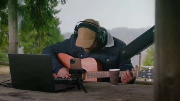 戴著耳机的白人会唱歌 在山上的木制凉亭里弹奏吉他 男性音乐家使用笔记本电脑 三脚架电话和专业话筒在室外创作和记录音乐或歌曲 — 图库视频影像