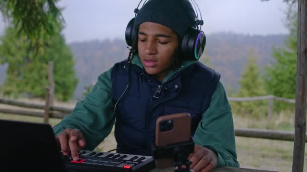 ヘッドフォンの若いミュージシャンは 屋外でMidiコントローラーを演じています アフリカ系アメリカ人のティーンエイジャーは 三脚のラップトップと携帯電話を使用して 山の木製のガゼボに座っている音楽を作成し 記録します スローモーション — ストック動画