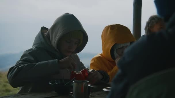 青少年男女坐在桌旁 走完长途跋涉后吃着饭 孩子们从袋子里吃远足的食物 并与其他游客一起在加泽博休息 登山探险期间的远足者 — 图库视频影像