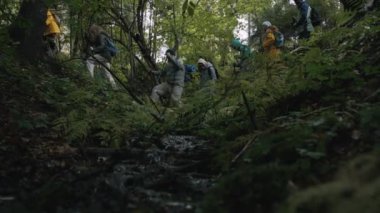 Sırt çantalı ve yürüyüş direkli çok ırklı turist grubu patika boyunca yürüyor. Dağlarda gezinti meraklıları. Güzel bir ormanda temiz bir dere akar. Ağır çekim. Düşük açılı çekim.