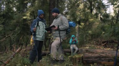 İki çok ırklı turist ormanda tablet bilgisayar kullanarak patikaları tartışıyor. Sırt çantalı ve yürüyüş direkli gezginler yürüyüş sırasında dinlenmek için dururlar. Doğa keşfi ve turizm kavramı. Yavaş çekim.