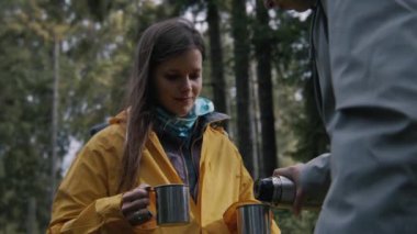 Dişi turist iki metal bardağı elinde tutarken, diğer gezgin termostan sıcak çay dolduruyor. Yürüyüş arkadaşları ormanda kamp yapmak için mola verdiler. Doğa keşfi ve turizm konsepti. Yavaş çekim.